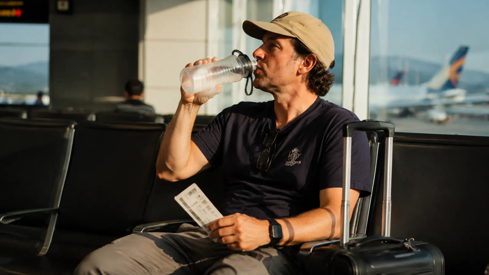 Traveler drinking from a collapsible water bottle while waiting at a gate with a rolling suitcase beside them